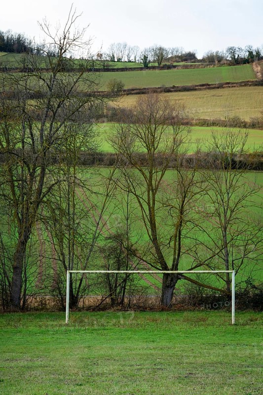 A serene countryside scene features two football goalposts set on a grassy field, bordered by leafless trees and lush green rolling hills, France