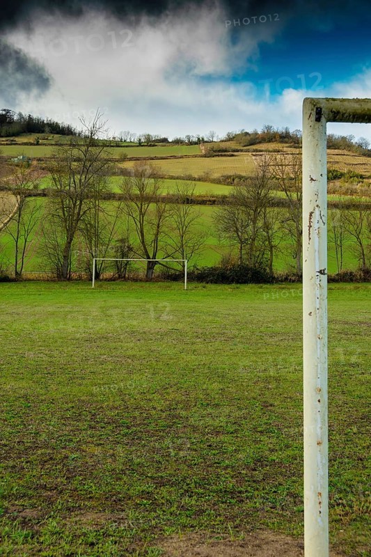 A football field is set against rolling hills and trees. The goalpost stands tall, framed by the vibrant greenery and dramatic clouds above, France