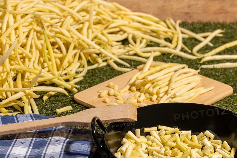Yellow runner beans next to a cooking pot with sliced beans, with a wooden spoon on top