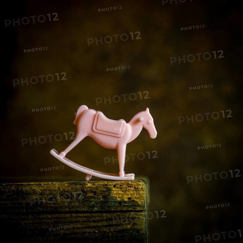 A small pink rocking horse toy sits on a weathered wooden shelf, gently illuminated in low light. The pastel color against the dark background evokes memories of childhood innocence and playfulness