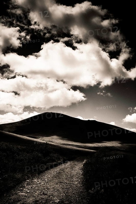 A winding path leads towards a dark mountain silhouette against a dramatic sky filled with billowing clouds. Sancy Massif, Puy de Dome, Auvergne, France
