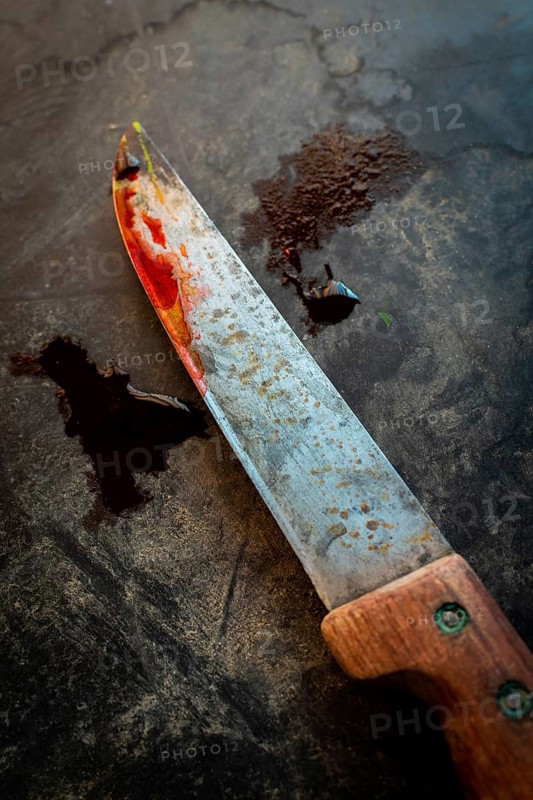 A worn knife with a rusty blade and wooden handle rests on a dark, textured surface. Traces of red stains hint at recent heavy use, sparking curiosity and caution