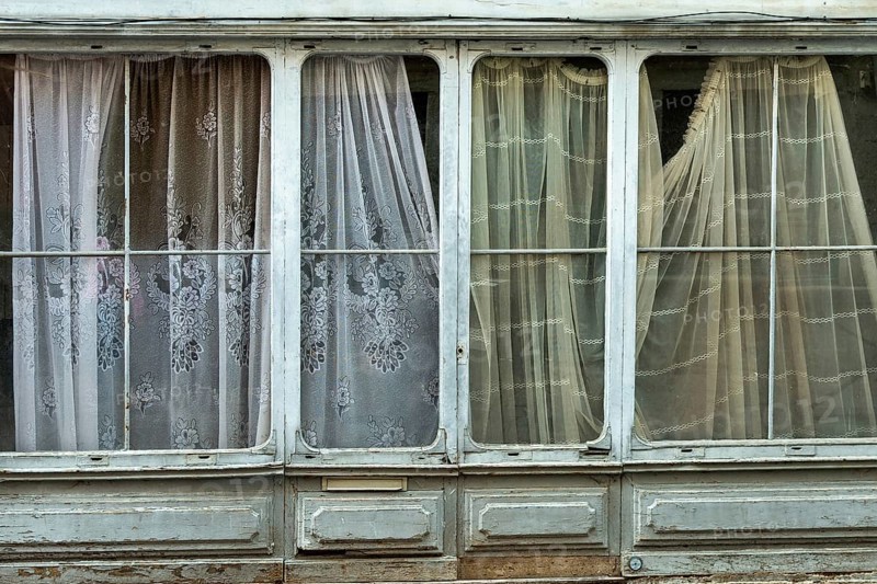 Light streams gently through patterned curtains hanging in a historic building, creating an atmosphere of calm and nostalgia. The weathered window frames hint at age and history. France