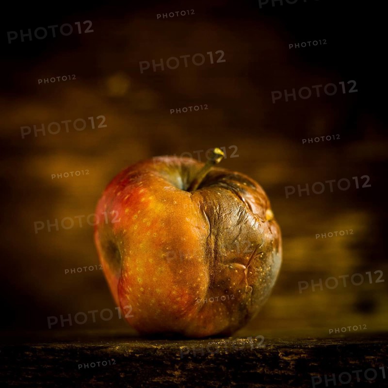 A decaying apple sits on a wooden surface, showcasing its wrinkled skin and faded color against a softly lit, dark background. This scene evokes a sense of time passing and decay