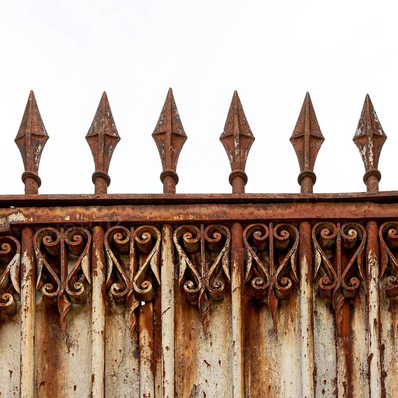 An old, weathered wrought iron fence features intricate scrollwork and pointed spikes atop its structure, showing signs of rust and age against a cloudy sky backdrop. France
