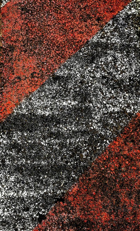 A close-up view of a textured pavement featuring bold diagonal stripes in red and black, showcasing worn surfaces and contrasting colors under midday sunlight