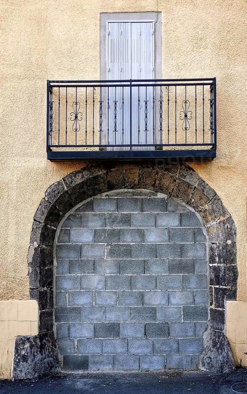A metal-railed balcony stands over a blocked stone entrance on a textured yellow building, creating a striking architectural feature in an urban area