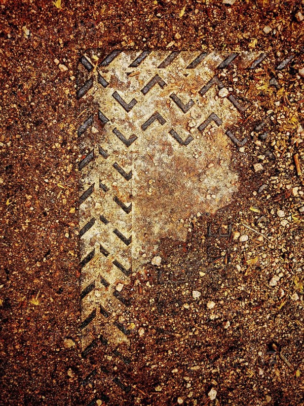 A worn metal cover is partially submerged in dirt and gravel, showcasing intricate patterns from erosion and weathering, highlighting nature's impact on human-made materials
