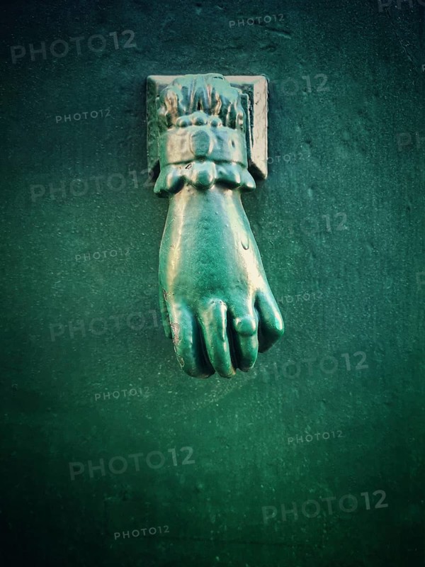 A unique green hand-shaped door knocker adorns a textured dark wall, adding a charming historical touch to the location. Its intricate design draws the eye and invites curiosity