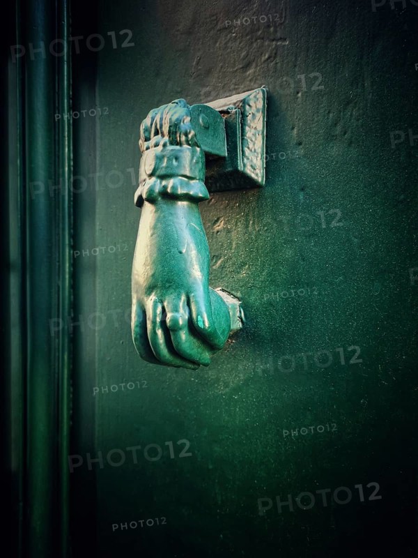 A distinctive bronze hand-shaped door knocker is mounted on a teal door, reflecting intricate craftsmanship while complementing the historic architecture of the building