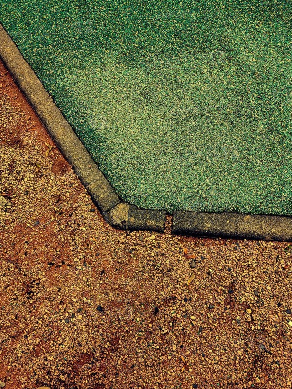 This detailed view captures the unique transition of a playground surface from vibrant green rubber to brown dirt during midday