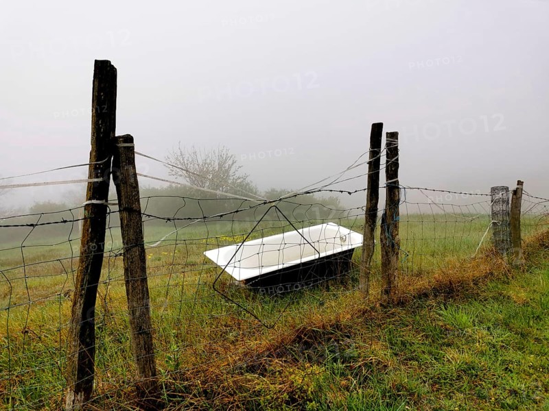 A rusted bathtub rests against a weathered fence, partially hidden by thick fog in a serene rural area during early morning. The mist enhances the peaceful atmosphere. france
