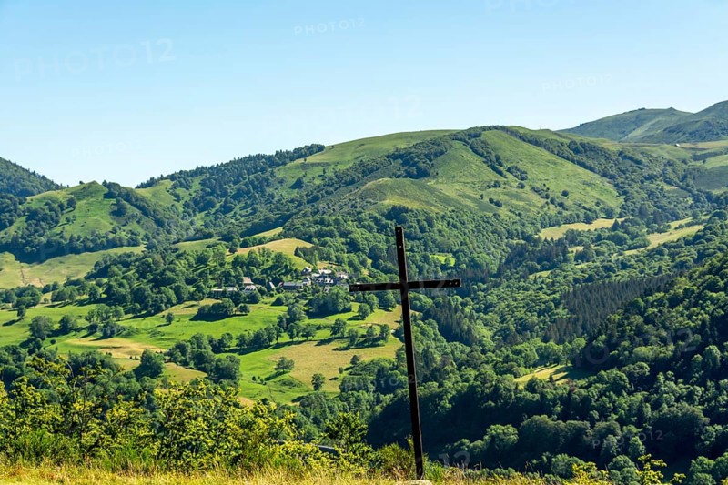 A wooden cross stands prominently against a picturesque landscape of rolling hills and vibrant greenery. The tranquil setting suggests a serene connection with nature, offering breathtaking views. Cezallier. Puy de Dome. Auvergne Rhone Alpes. France