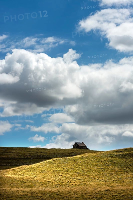 Ruined stone cottage stands alone on a grassy hillside under a partly cloudy sky in a remote countryside setting. Cezallier. Puy de Dome. Auvergne Rhone Alpes. France