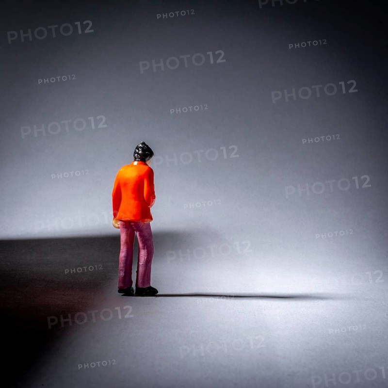 A miniature figure dressed in an orange jacket stands still on a smooth surface, surrounded by shadows while illuminated by focused light, creating a dramatic effect