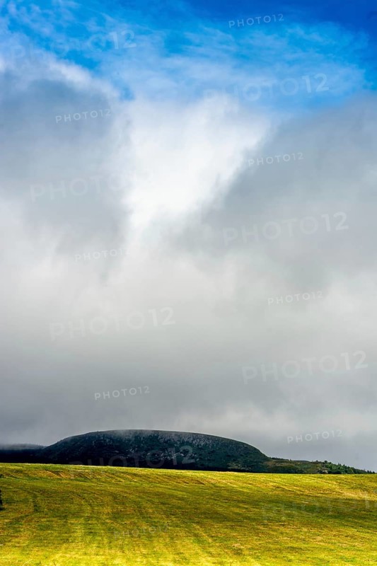A bright yellow field stretches across the landscape, illuminated by sunlight that peeks through the clouds. A dark hill looms in the distance, enhancing the serene atmosphere. Puy de Dome. Auvergne Rhone Alpes. France