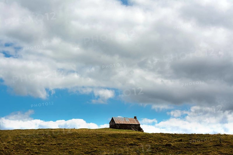 A small cabin sits alone atop a grassy hill, surrounded by wide-open space. The sky is filled with fluffy clouds, creating a serene atmosphere in this rural landscape. Cezallier. Puy de Dome. Auvergne Rhone Alpes. France