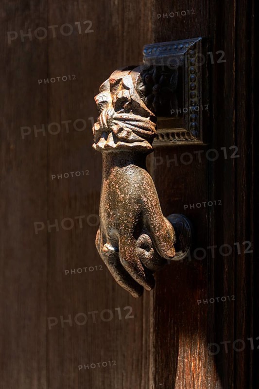 A detailed bronze hand-shaped door knocker is affixed to a rustic wooden door, showcasing intricate design and historical craftsmanship under natural light