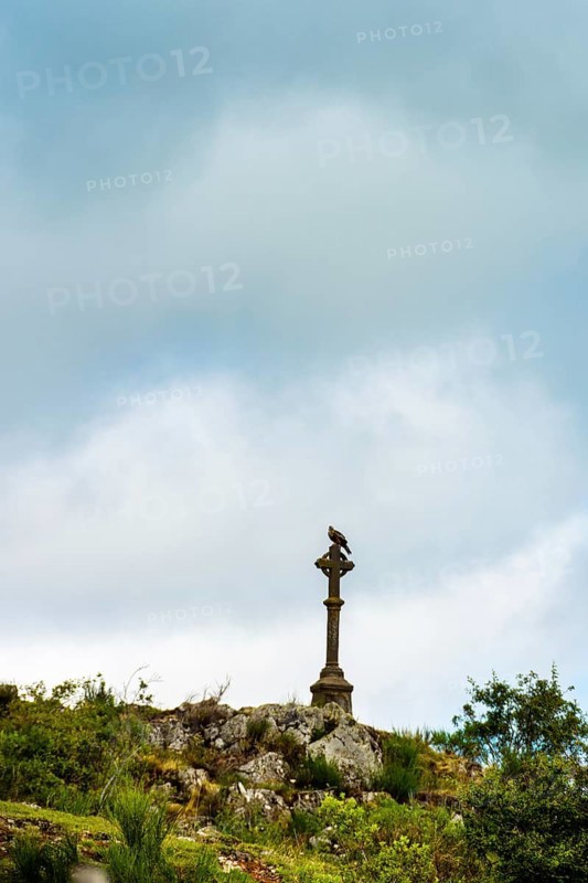 A stone cross towers over rocky ground, surrounded by greenery. Dark clouds loom overhead, creating a dramatic backdrop that enhances the peacefulness of the area. Puy de Dome. Auvergne Rhone Alpes. France