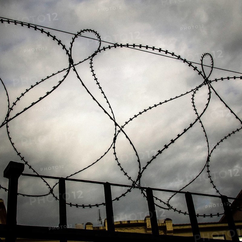 Rusty barbed wire coils twist against a dark and turbulent sky, creating a sense of confinement and unease. The storm clouds loom over a distant structure, emphasizing isolation
