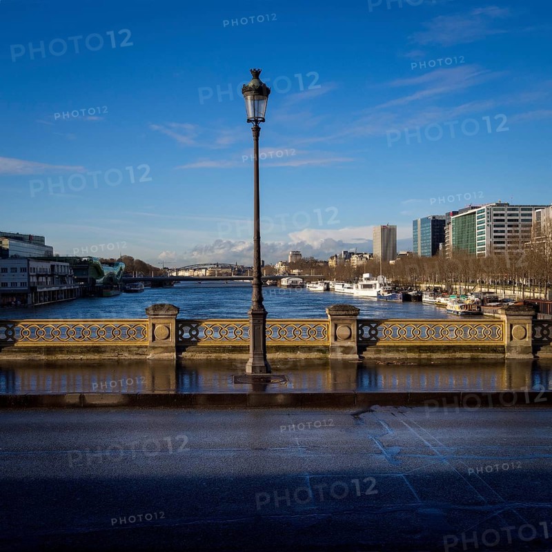 A street light stands tall on a bridge, casting light on the path as the serene Seine River flows beneath. Nearby buildings reflect a calm sky, showcasing urban life in Paris. France