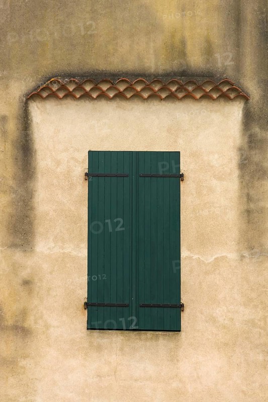 The facade features a pair of blue-green wooden shutters on a weathered wall, showcasing texture and subtle architectural details highlighting the age and character of the house. France