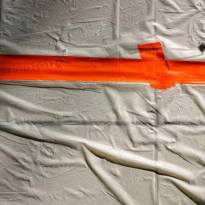 Bright orange tape stretches across a wrinkled white surface, creating a clear boundary. This setup suggests preparation for an artistic process or craft activity in a studio