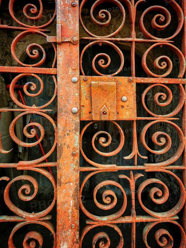 A weathered iron gate showcases detailed swirling patterns, highlighting years of exposure to the elements. The rust adds character, framing a glimpse of the aged building behind