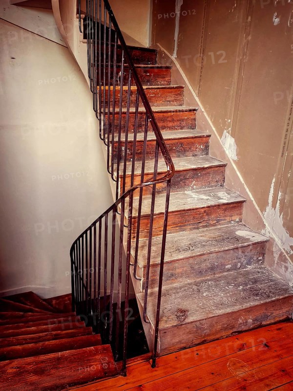 A winding staircase constructed from rustic wood features elegant iron railings, set in a partially renovated interior with unfinished walls. Light filters in, highlighting textures