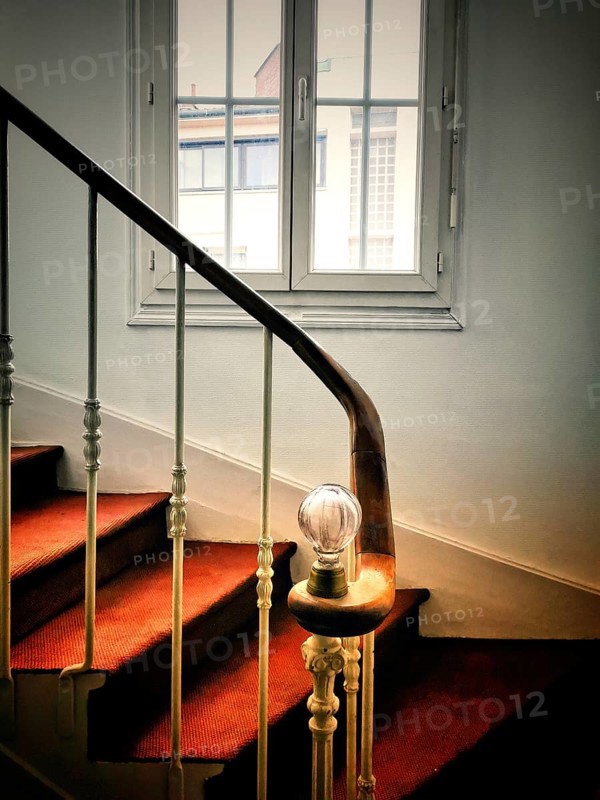 A staircase is gracefully illuminated by a soft light bulb on the railing, leading upward with a carpeted surface and natural light entering through a nearby window