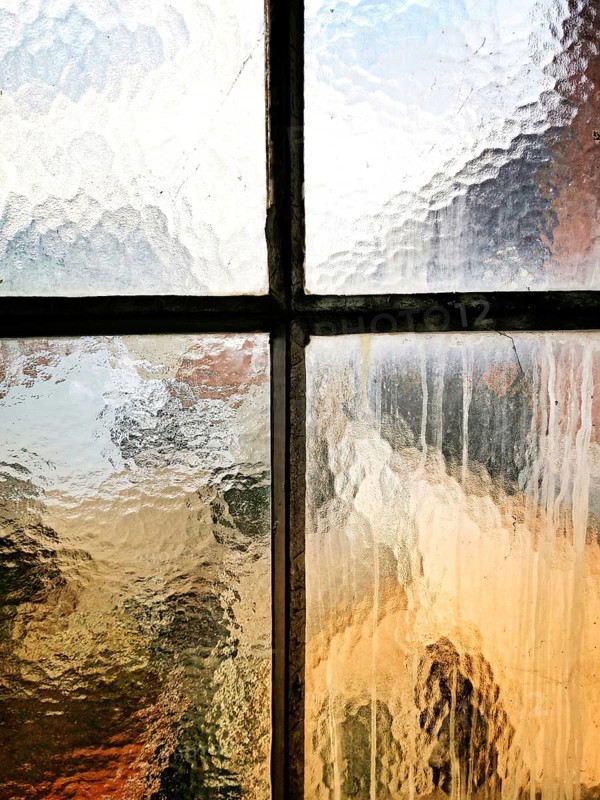 A textured glass window shows raindrops trickling down. The blurred outlines of an urban landscape and warm colors create a cozy, serene atmosphere