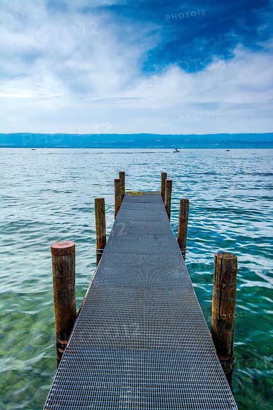 A sturdy jetty extends into the serene waters of Lake Geneva, surrounded by mesmerizing blue hues and a tranquil atmosphere. Perfectly captures the beauty of Haute-Savoie on a peaceful day, Auvergne Rhone Alpes, France