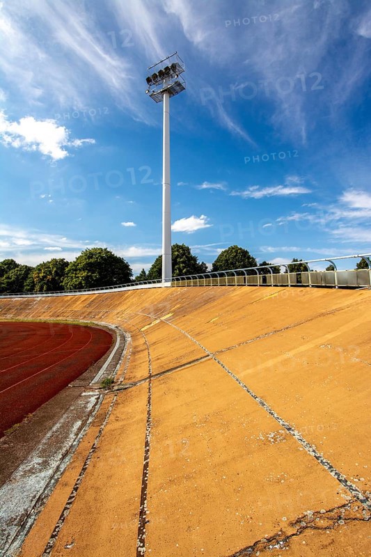 Bright daylight reveals an empty sports stadium with a vibrant red track. The tall floodlights, overgrown with neglect, indicate years without activity. The clear blue sky adds to the desolation. France