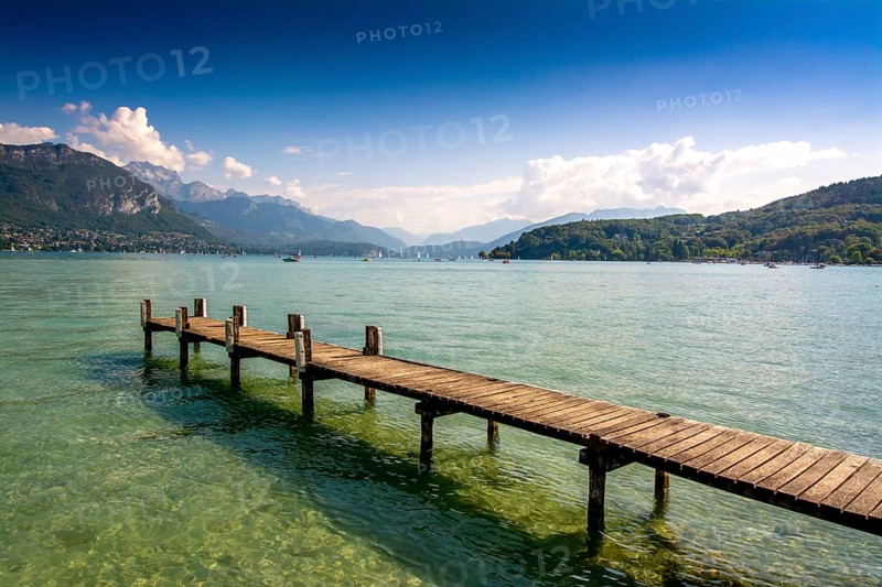 Clear skies reflect on the calm waters of Annecy lake, while a wooden pontoon invites visitors to enjoy the breathtaking mountain scenery in Haute-Savoie, France