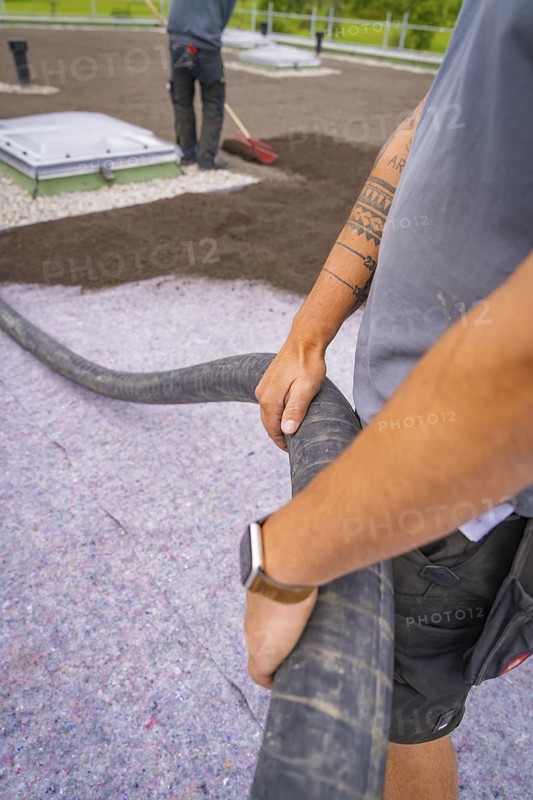 Worker holds a hose with visible tattoos and uses construction clothing, roof greening, house construction, climate neutral