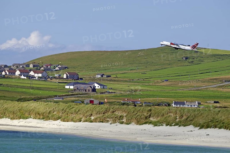 An aeroplane takes off over wide meadows and a coastal village with sea views, Sumburgh Head, Shetland Islands, Scotland, United Kingdom