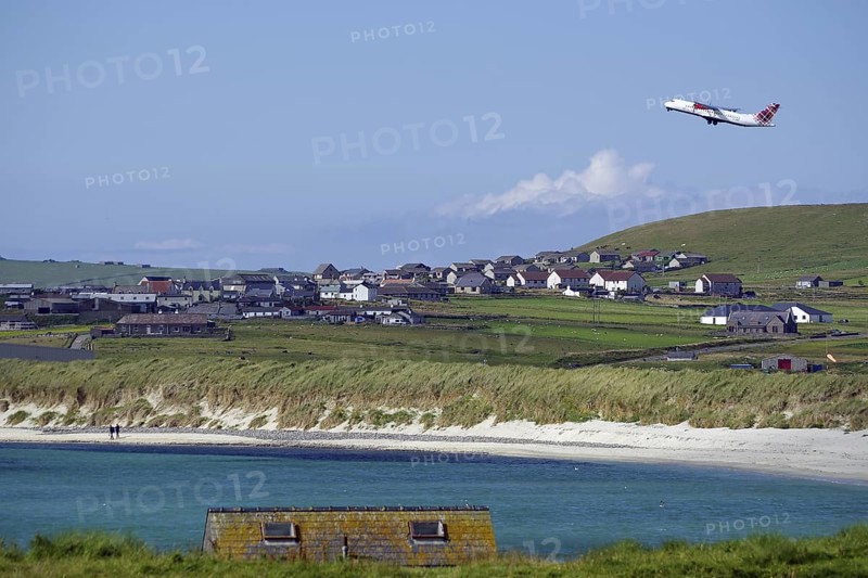 An aeroplane takes off near a rural village with beach and meadows, Sumburgh Head, Shetland Islands, Scotland, United Kingdom