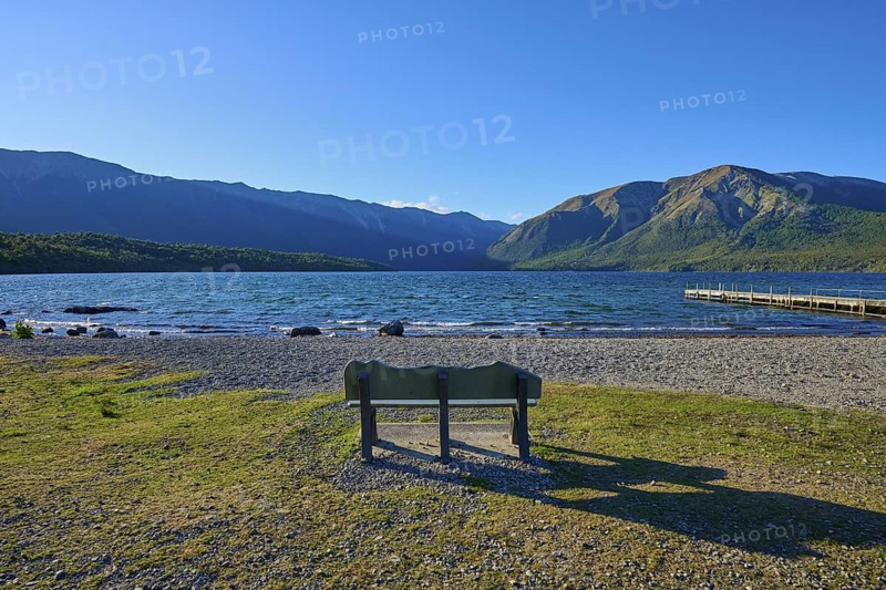 A park bench on the shore of a lake with a view of the surrounding mountains and a boardwalk under a clear blue sky, summer, Lake Rotoiti, Saint Arnaud, Tasman Region, South Island, New Zealand