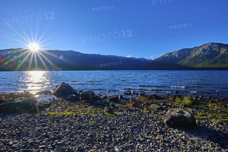 Sunlight dazzles over the horizon as it hits the tranquil lake and surrounding mountains, summer, Lake Rotoiti, Saint Arnaud, Tasman Region, South Island, New Zealand