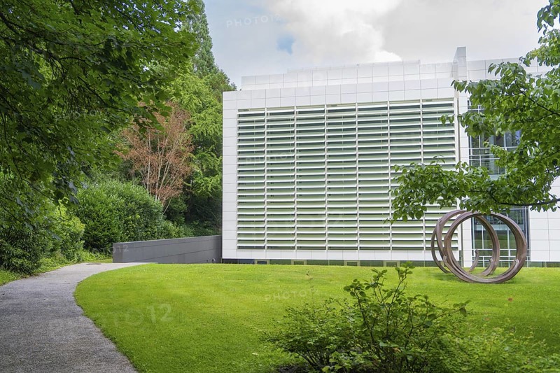 Modern building with a green façade, surrounded by nature and a sculpture in front. A path leads through the grounds, Baden Baden, Germany