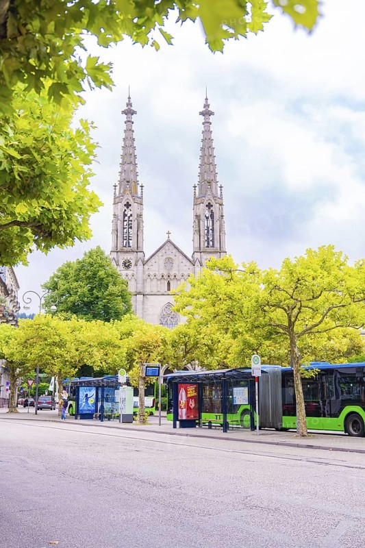 Gothic church with towers behind trees, buses stop on a busy street, Baden Baden, Germany