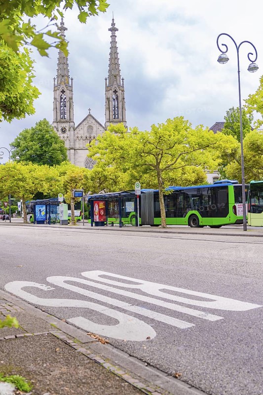 An urban street with buses, flanked by trees, gothic church towers visible in the background, Baden Baden, Germany