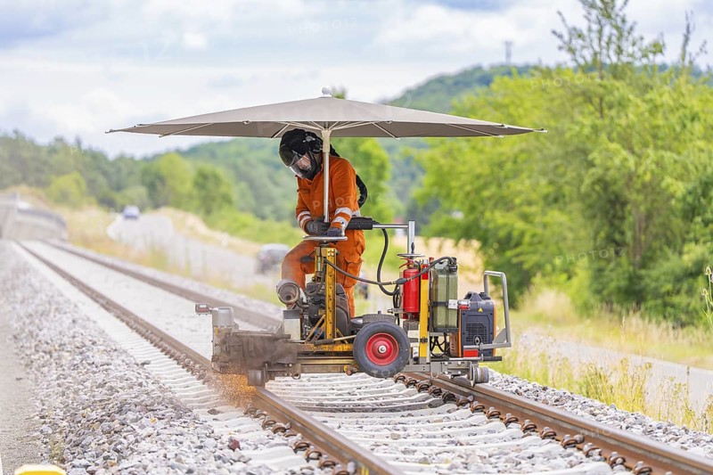 Worker in orange overalls operating a machine on railway tracks, protected by a large umbrella, grinding work on the Hermann Hesse railway, Ostelsheim, district of Calw, Germany