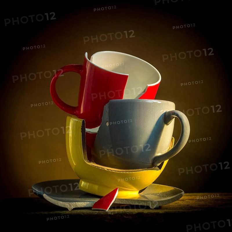 A collection of broken tableware is displayed, featuring a red mug and plate alongside other cracked cups. The setting is dimly lit, enhancing the fragile nature of the pottery
