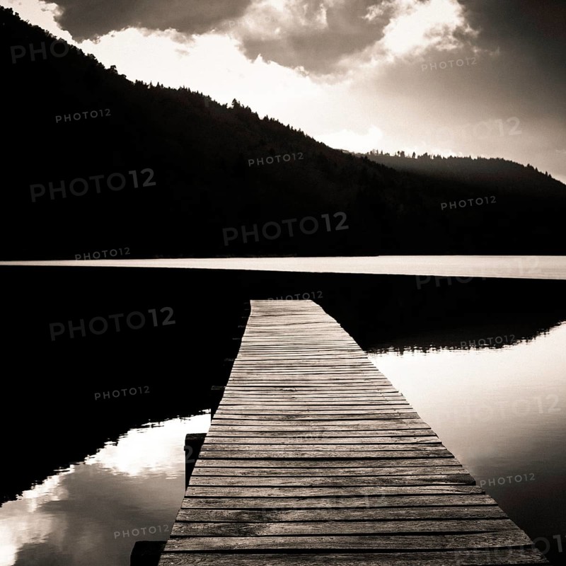 A tranquil wooden pier stretches out over the calm waters of Lake Chambon, surrounded by lush hills in Auvergne, France. This idyllic setting invites peace and relaxation, Puy de Dome, Auvergne Rhone Alpes, France