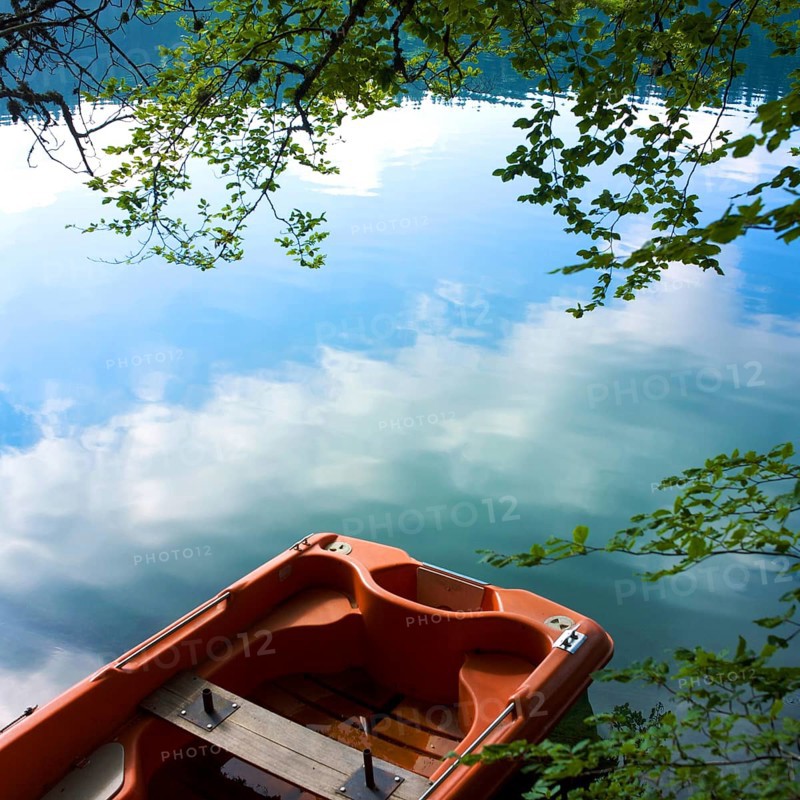 A small boat rests quietly on the tranquil waters of Lake Pavin, surrounded by lush greenery. The clear reflections of clouds and trees create a serene atmosphere of natural beauty, Puy de Dome, Auvergne Rhone Alpes, France