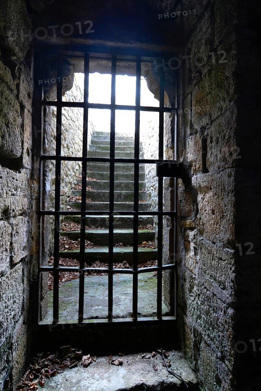 A medieval dungeon in France features thick stone walls and a barred window revealing illuminated stone steps. Leaves cover the floor, enhancing the haunting atmosphere