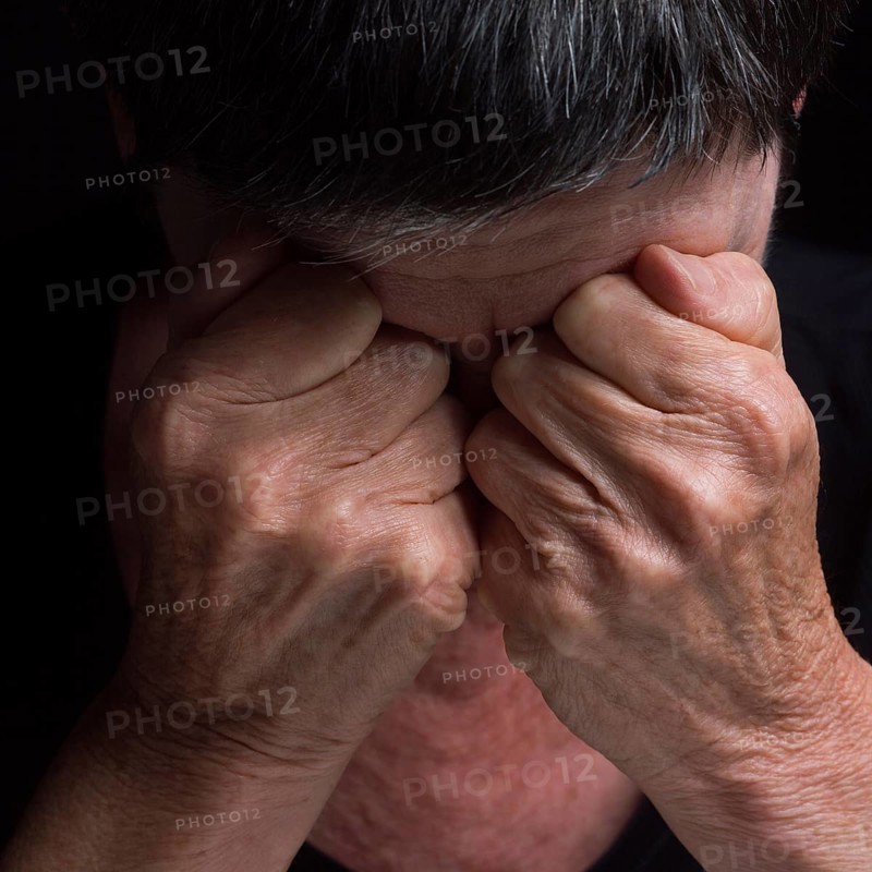 A person sits with their head bowed, hands clasped tightly over their eyes, conveying deep emotion and turmoil. The setting is dark, emphasizing feelings of distress and introspection