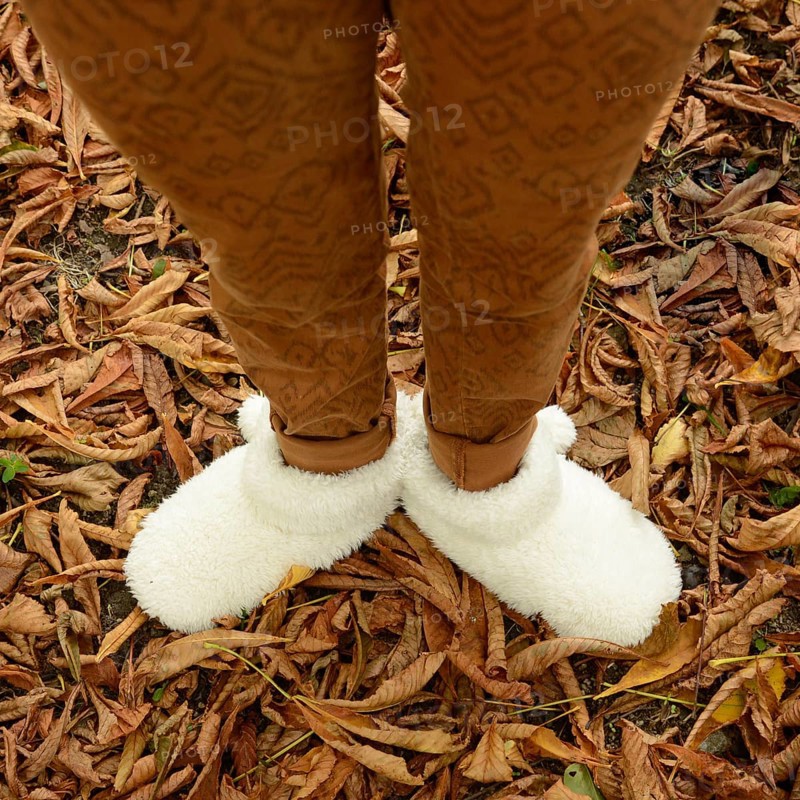 A young woman stands among fallen leaves, wearing warm, fluffy shoes that provide comfort. The autumn landscape reflects vibrant colors, enhancing the cozy atmosphere of the season