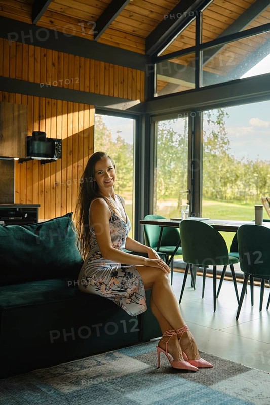 Happy woman in a floral dress sits on a couch in a sunlit room with large windows and contemporary decor
